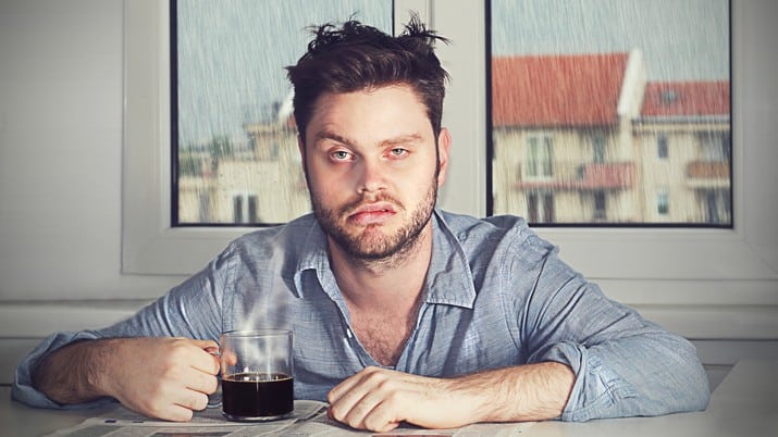 A man slumps crankily over his morning coffee as it pours with rain outside.