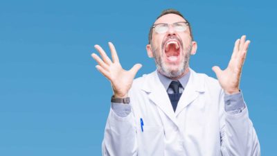 A man wearing a white coat holds his hands up and mouth open with joy.