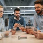 A group of people sit around a table playing cards in a work office style setting.