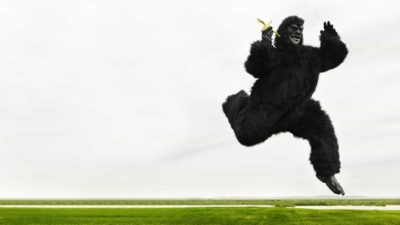 A person in a gorilla suit leaps really high holding a banana, nearly doing the splits.