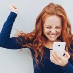 A woman with strawberry blonde hair has a huge smile on her face and fist pumps the air having seen good news on her phone.