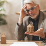 A man sits at a desk holding a small replica house in his hand, upset at the sale of his property.