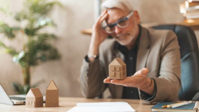 A man sits at a desk holding a small replica house in his hand, upset at the sale of his property.