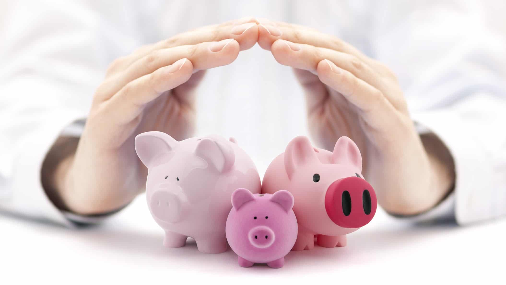 A person holds their hands over three piggy banks, protecting and shielding their money and investments.