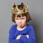 A little girl wearing a gold crown sulks and pokes her tongue out.