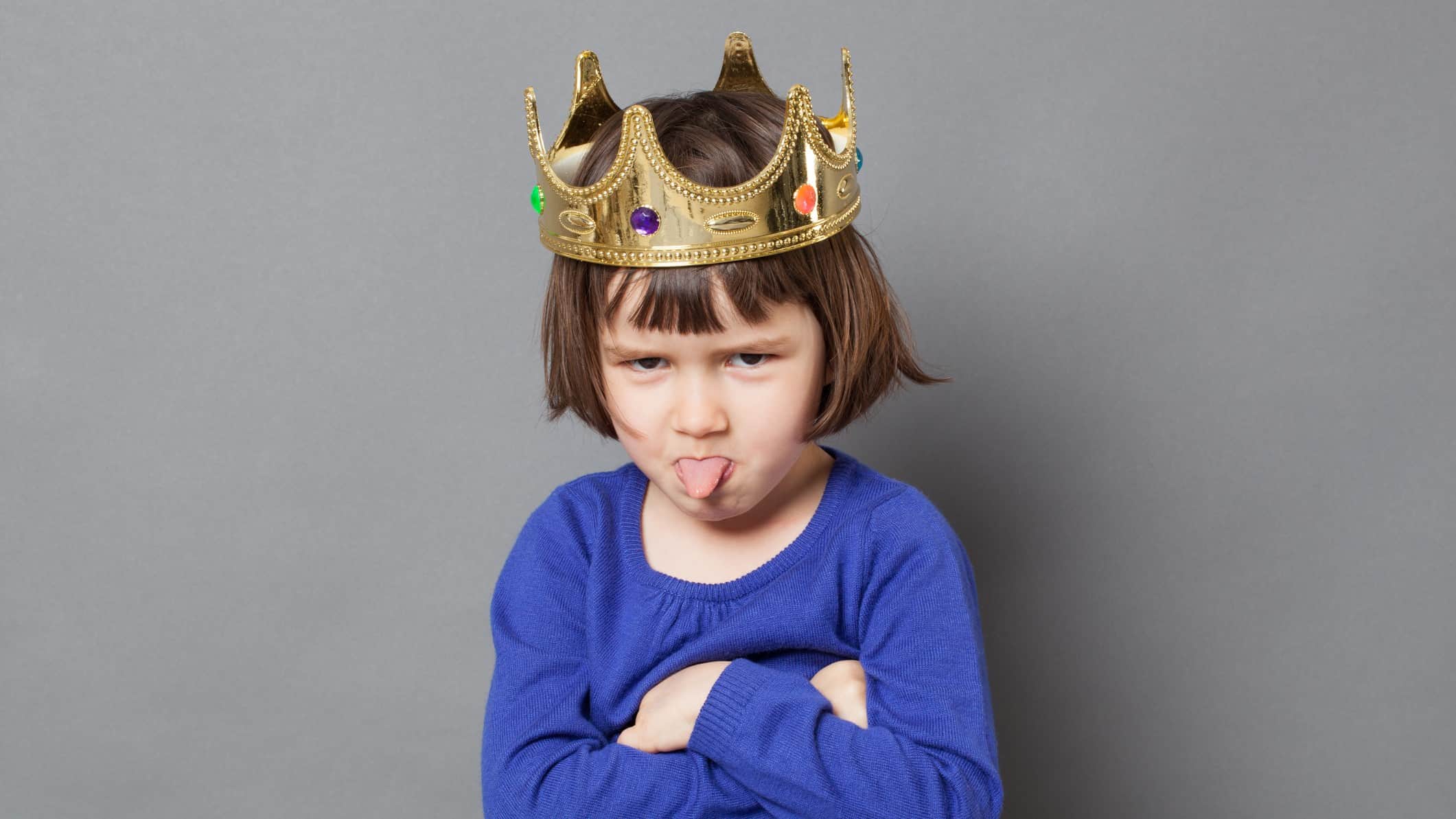 A little girl wearing a gold crown sulks and pokes her tongue out.