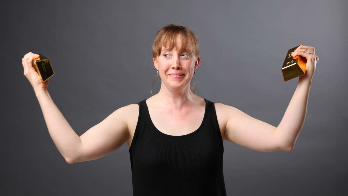 A woman holds a gold bullion in each hand, arms out showing her muscles with an incredulous look on her face.