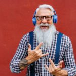 A trendy older hipster guy with a long white beard and headphones pulls rockstar hand sign with his hands.