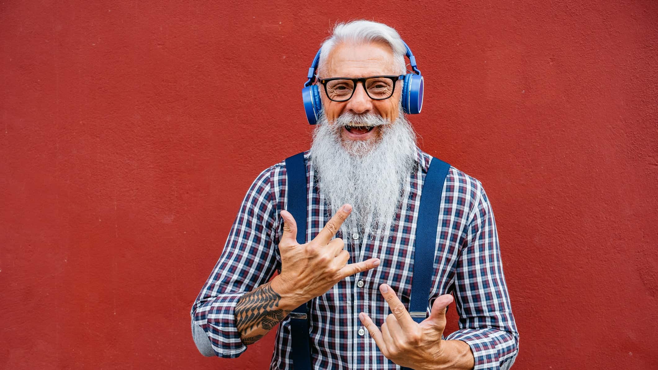 A trendy older hipster guy with a long white beard and headphones pulls rockstar hand sign with his hands.