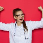 A scientist in a white coat and glasses puts her arms in the air in a sign of strength and success.