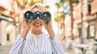 A woman standing on the street looks through binoculars.
