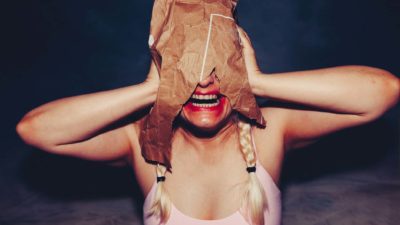 A woman with smeared lipstick and a paper bag on her head reveals her teeth in a half smile half scream.