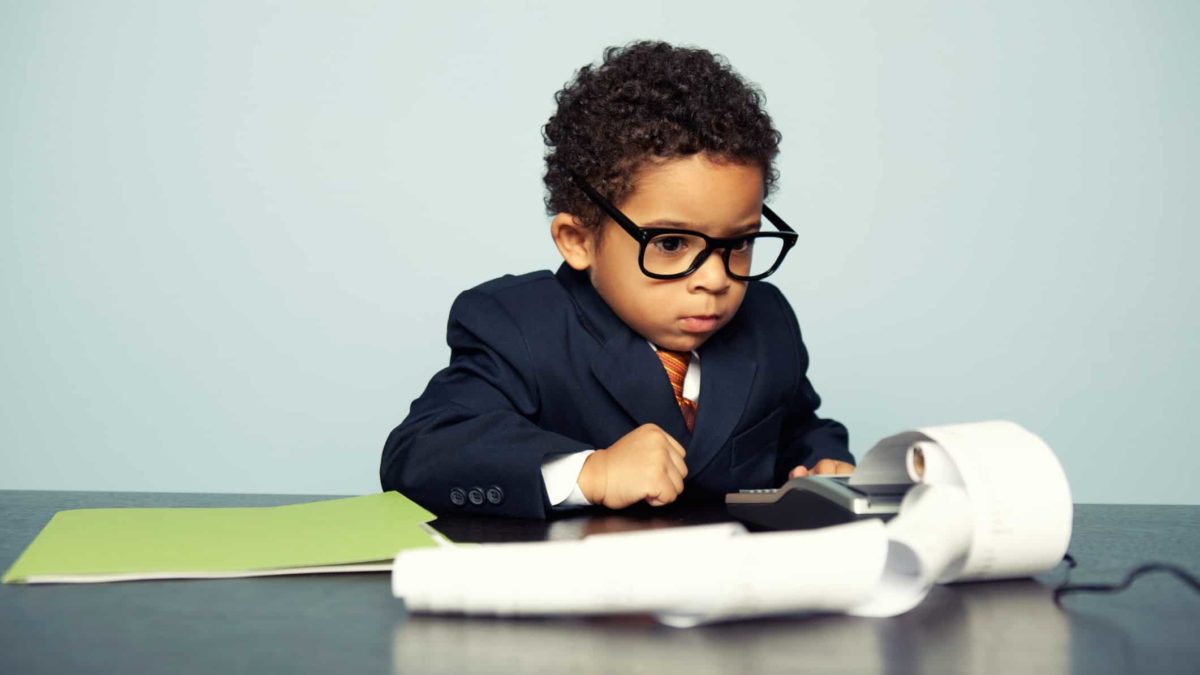 a cute little boy with curly hair wearing a business suit with a tie and too big glasses looks intently at an old fashioned business calculator with a scroll of paper spilling onto his desktop.