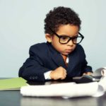 A cute little boy with curly hair wearing a business suit with a tie and big glasses looks intently at an old fashioned business calculator with a scroll of paper spilling onto his desktop.