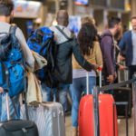 a crowd of people at an airport stand, some in queues, others looking around, while all drag their bags on wheels beside them.