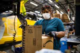 a warehouse worker wearing a face mask handles a cardboard box in an automated warehouse setting with equipment in the background.