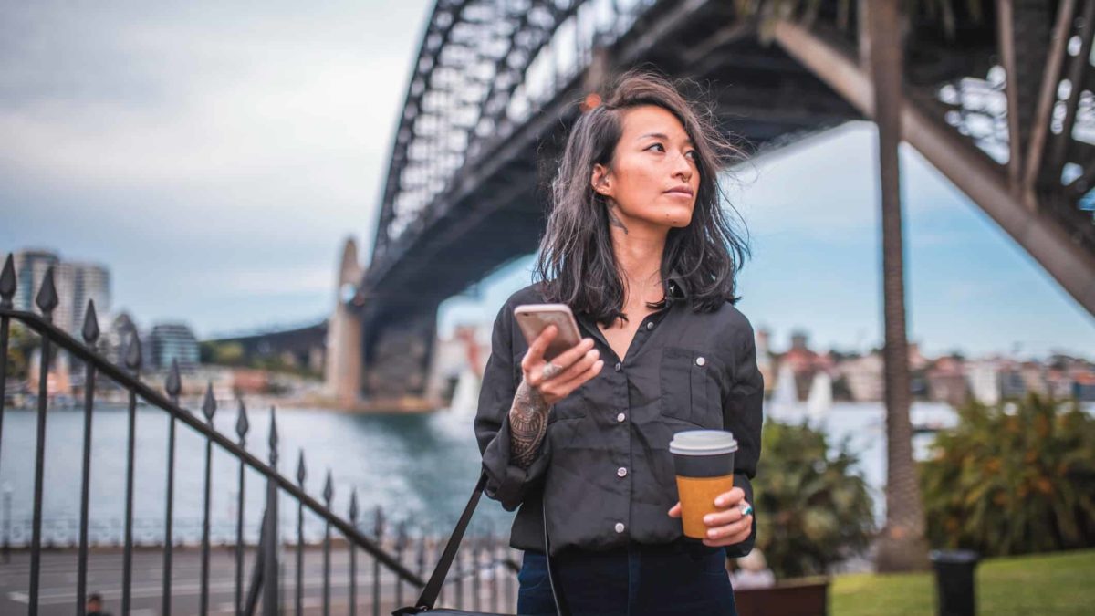 A woman wearing dark clothing and sporting a few tattoos and piercings holds a phone and a takeaway coffee cup as she strolls under the Sydney Harbour Bridge which looms in the background.