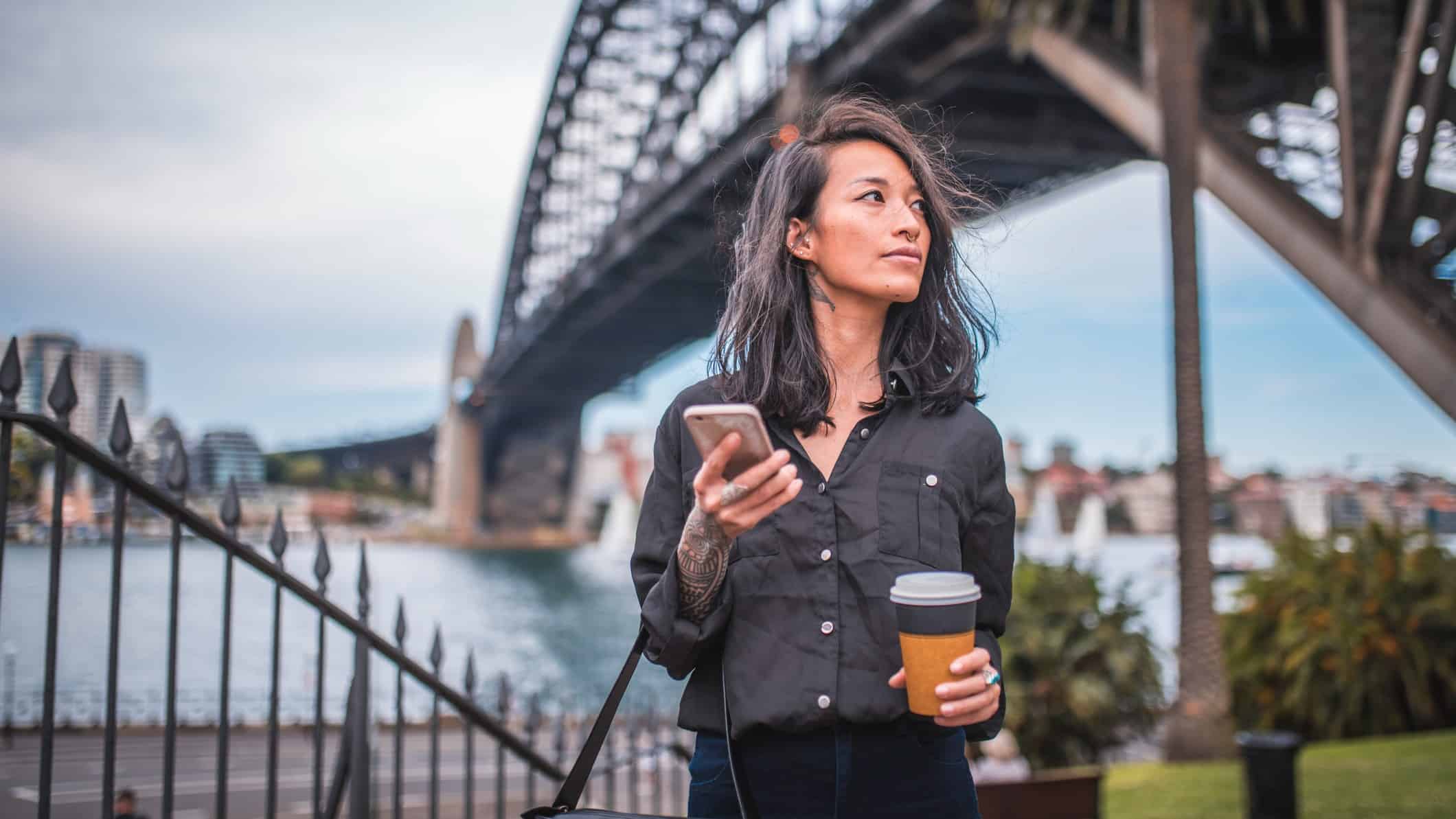 a woman wearing dark clothing and sporting a few tatoos and piercings holds a phone and a takeaway coffee cup as she strolls under the Sydney Harbour Bridge which looms in the background.