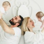 a couple in a bed hold a baby each up in the air, indicating they are the parents of twins. They look happy as they hold the babies aloft.