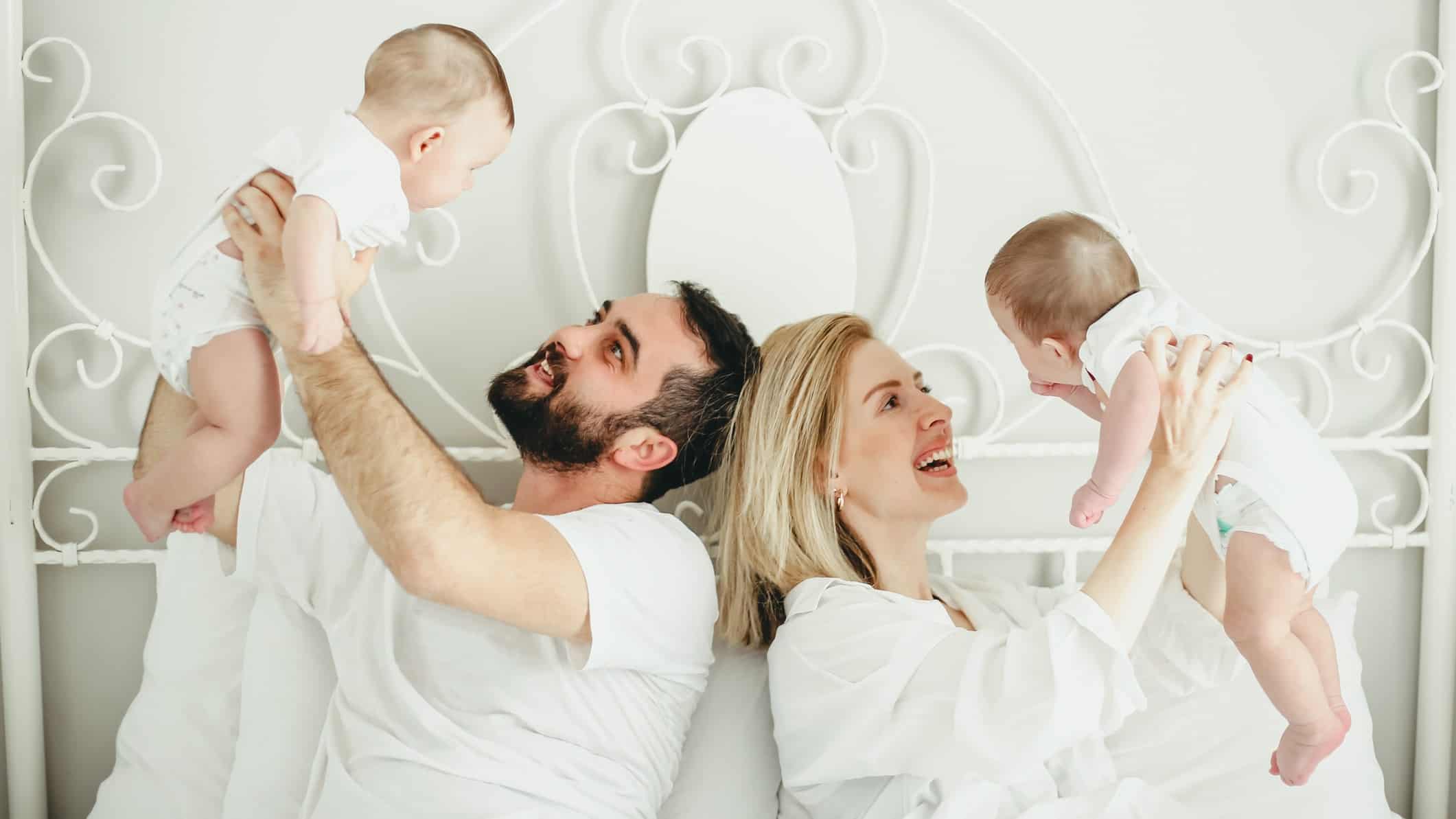 a couple in a bed hold a baby each up in the air, indicating they are the parents of twins. They look happy as they hold the babies aloft.