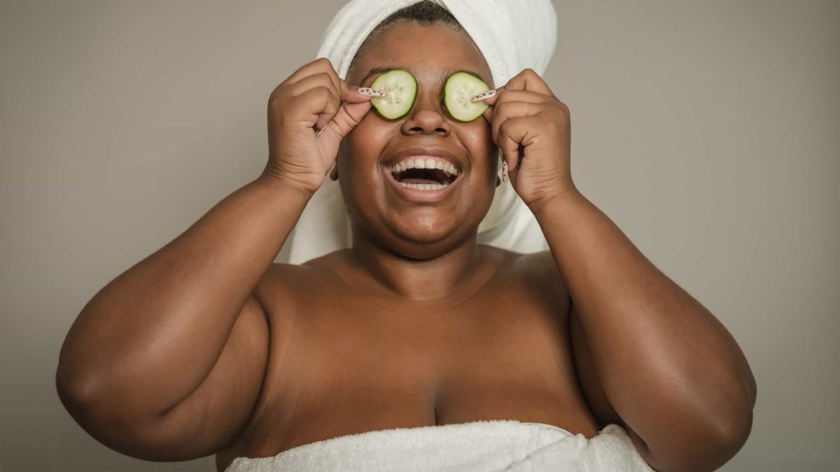 a happy woman wearing a white towel around her chest and another around her head laughs heartily while holding two slices of cucumber over her eyes as part of a beauty regime.