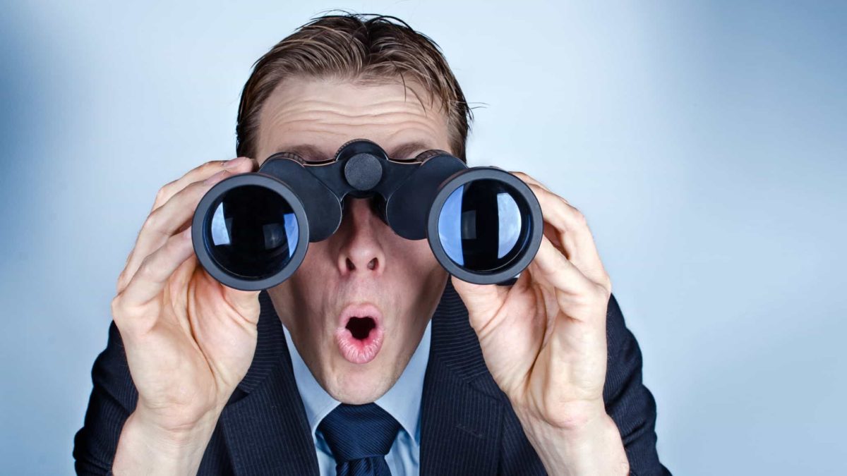 A man in a suit looks surprised as he looks through binoculars.