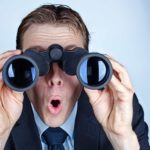 A man in a suit looks surprised as he looks through binoculars.