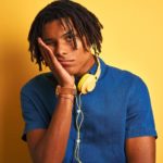 A young man wears headphones around his neck and holds his hand to his face as he leans into it with a sad, mournful look.