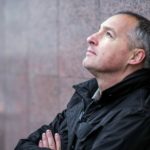 A concerned man leans against a brick wall looking up at the sky.