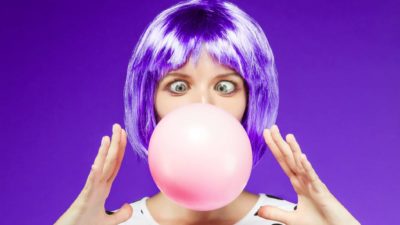 a woman with bright artificially coloured hair blows a large bubble gum bubble from her mouth with her eyes wide open and holding her hands either side of it.