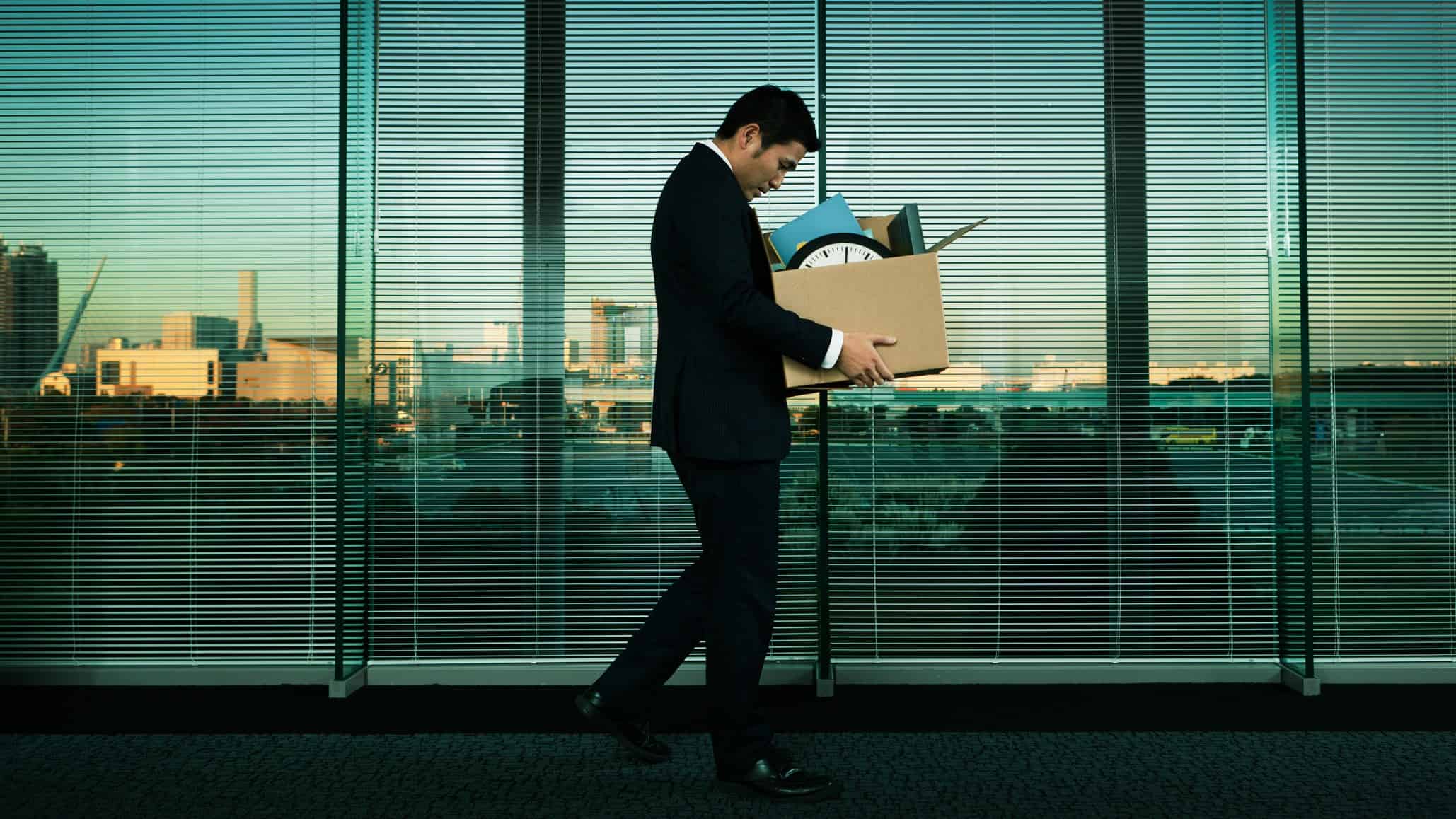 A man walks dejectedly with his belongings in a cardboard box against a background of office-style venetian blinds as though he has been giving his marching orders from his place of employment.
