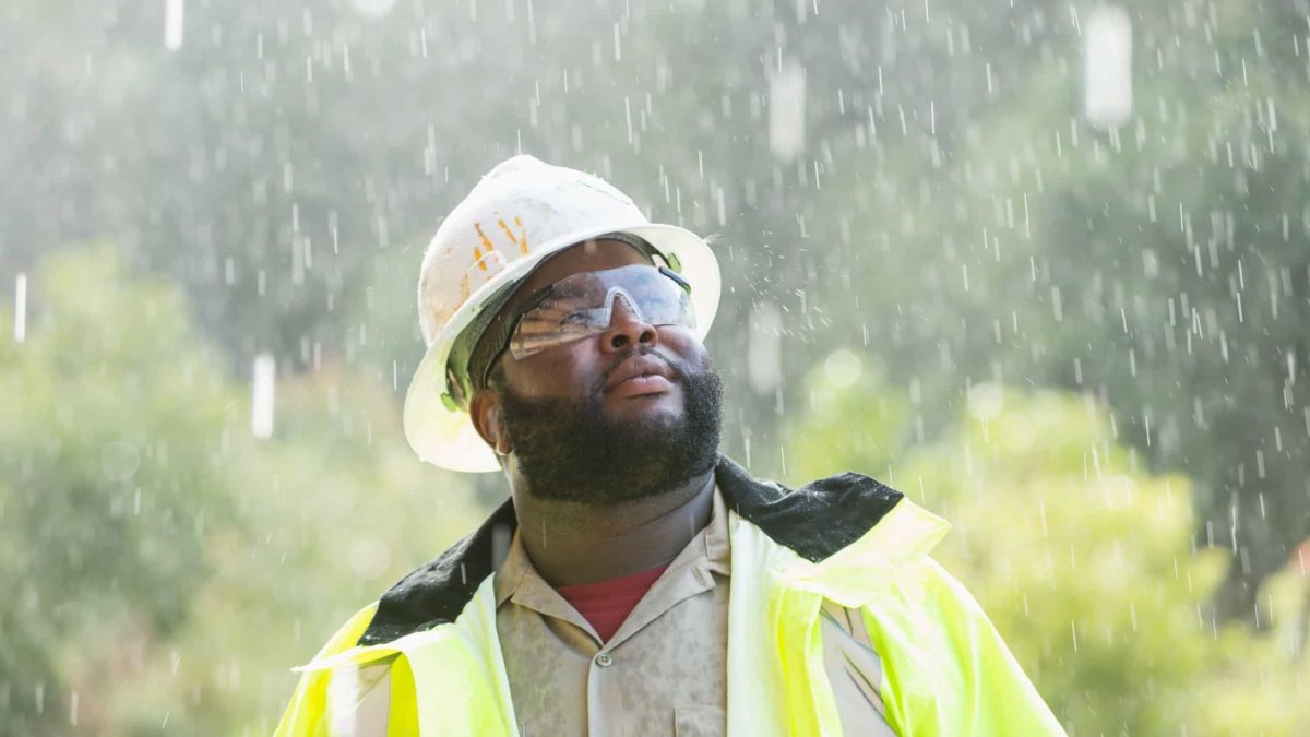 a construction worker in high visibility vest, hard hat and safety glasses looks up to the sky as rain falls and wets his clothing and face.