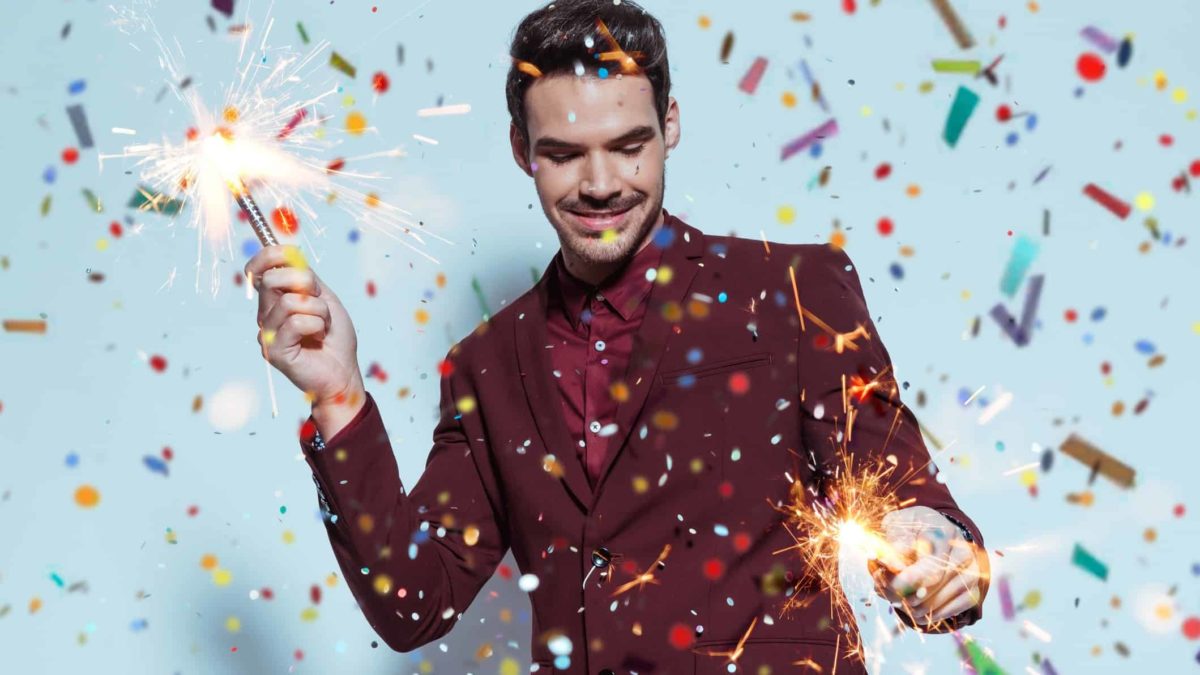 a man holds a firework sparkler in both hands as a shower of sparkly confetti falls from the sky around him as he smiles and closes his eyes in a celebratory scene.