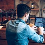 a man with his back facing the camera sits at a computer displaying a screen of code with an electric power contraption on the desk near him as he sits in concentration while appearing to mine cryptocurrency.