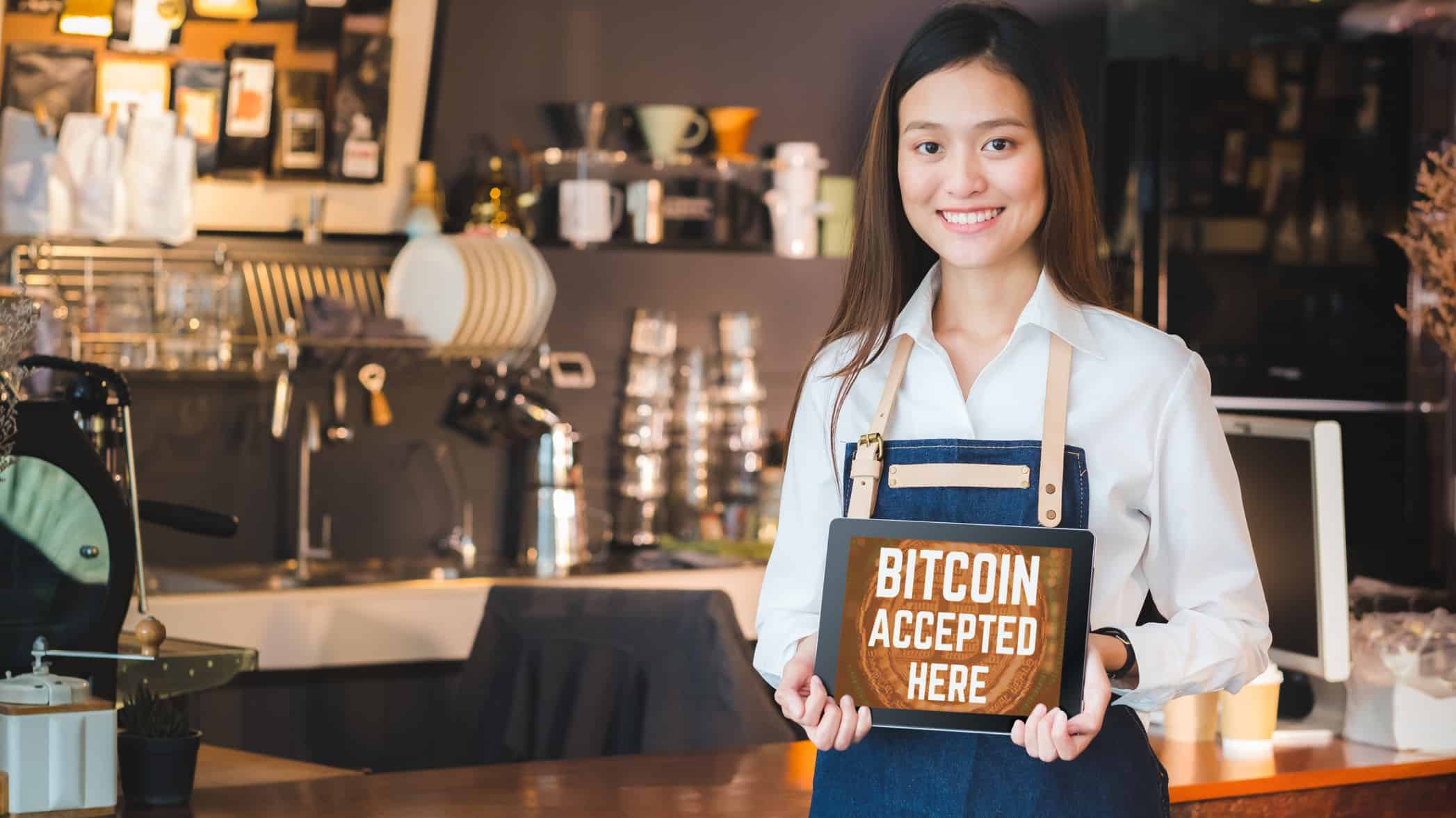 a smiling female cafe worker wearing an apron stands in front of an elaborate coffee machine and cafe set up with glasses and cups on shelves as she holds a sign that says 'Bitcoin Accepted Here' .