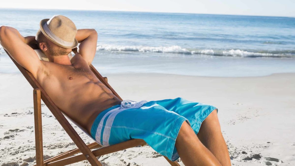 A man wearing only boardshorts stretches back on a deck chair with his arms behind his head and a hat pulled down over his face amid an idyllic beach background.
