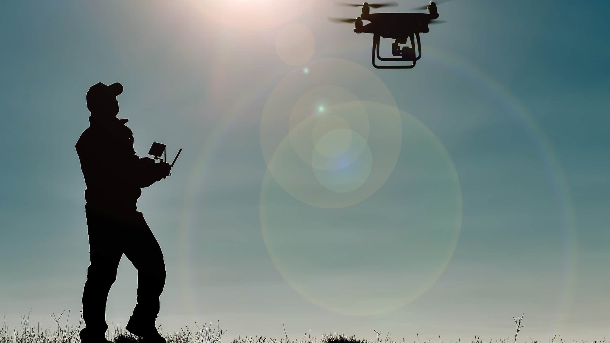 A silhouette shot of a man holding a control in his hands and watching as a drone hovers overhead with sunrays coming from the sky.