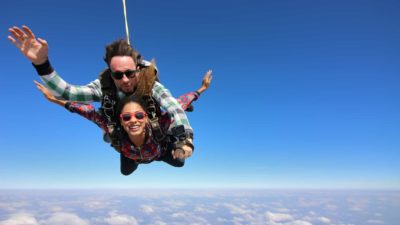 two smiling people, a man and a woman, raise a hand in a wave as they are tethered to each other while they skydive against a clear sky with a covering of clouds below before their parachute opens.