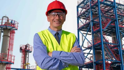 a gas worker with hard hat and high visibility vest stands cross armed and smiling in front of an elaborate steel structured gas plant.