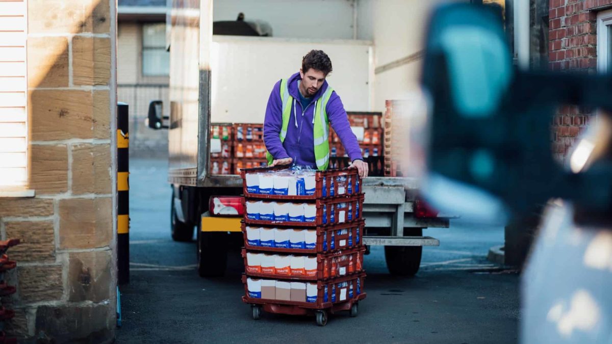 A man handles a pallet of grocerry goods stacked in rows in a warehouse area as though he is going to load it onto a trucl whose mirror can be seen in the foreground of the picture.