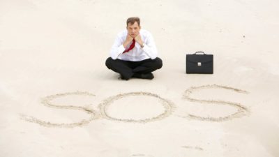 a businessman wearing tousers, a shirt and a tie sits cross legged on the sand in front of a sign that says SOS with his brief case beside him.