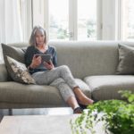 A woman sits amid a stylish home setting on a sofa with plush cushions with a coffee table and plant in the foreground while she peruses a tablet device.