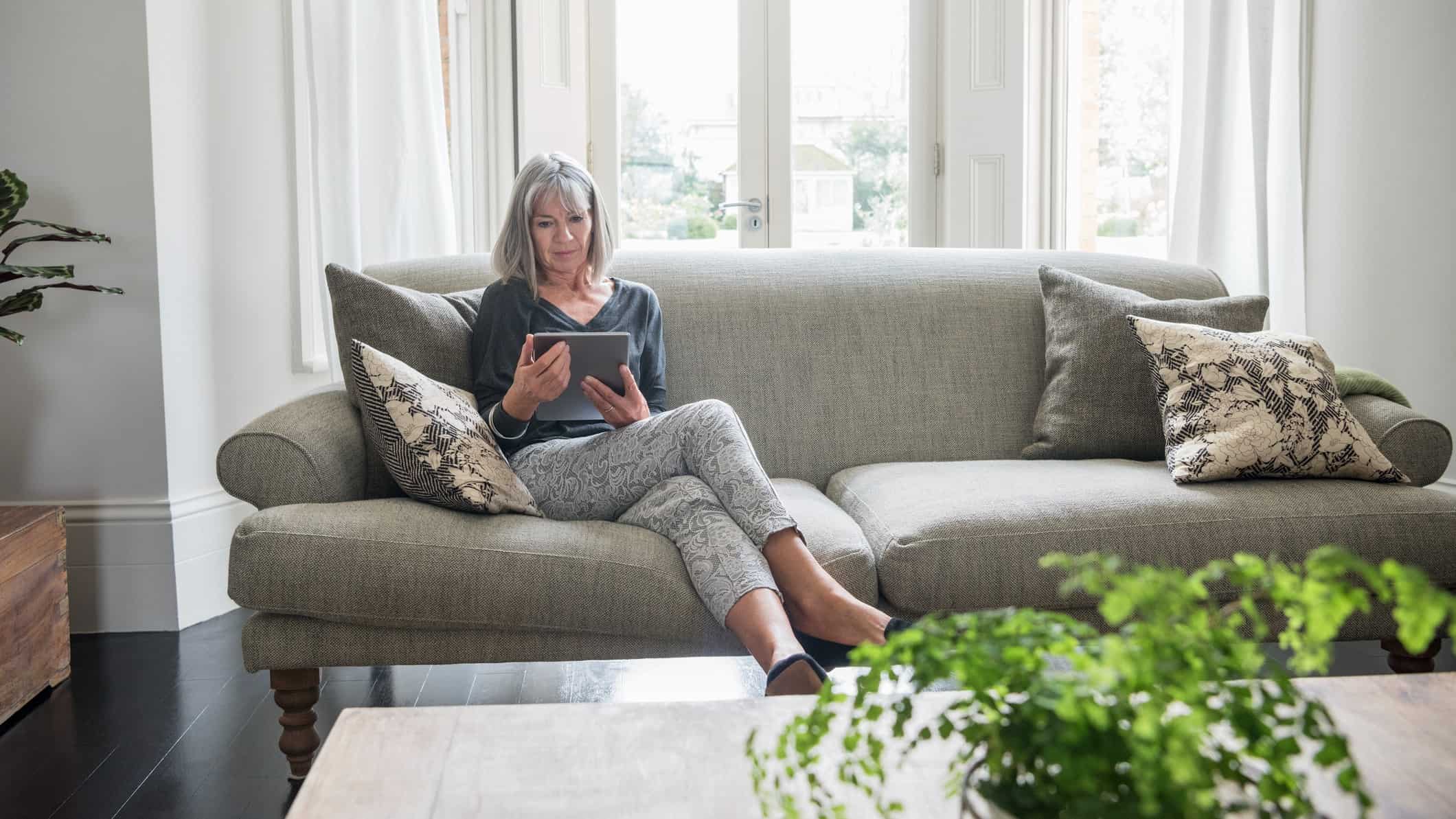 a woman sits amid a stylish home setting on a sofa with plush cushions with a coffee table and plant in the foreground while she peruses a tablet device.