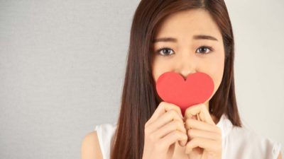 an attractive young woman with sad eyes holds a red paper love heart over her mouth as though she has been unlucky in love.