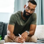 a man sits in a home environment on a sofa while writing in a book with a pen, a plant on the table nearby and curtains open in the background.