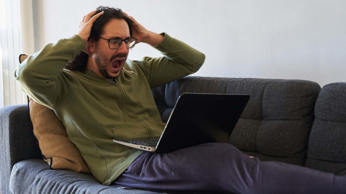 Stock market crash concept of young man screaming at laptop on the sofa.