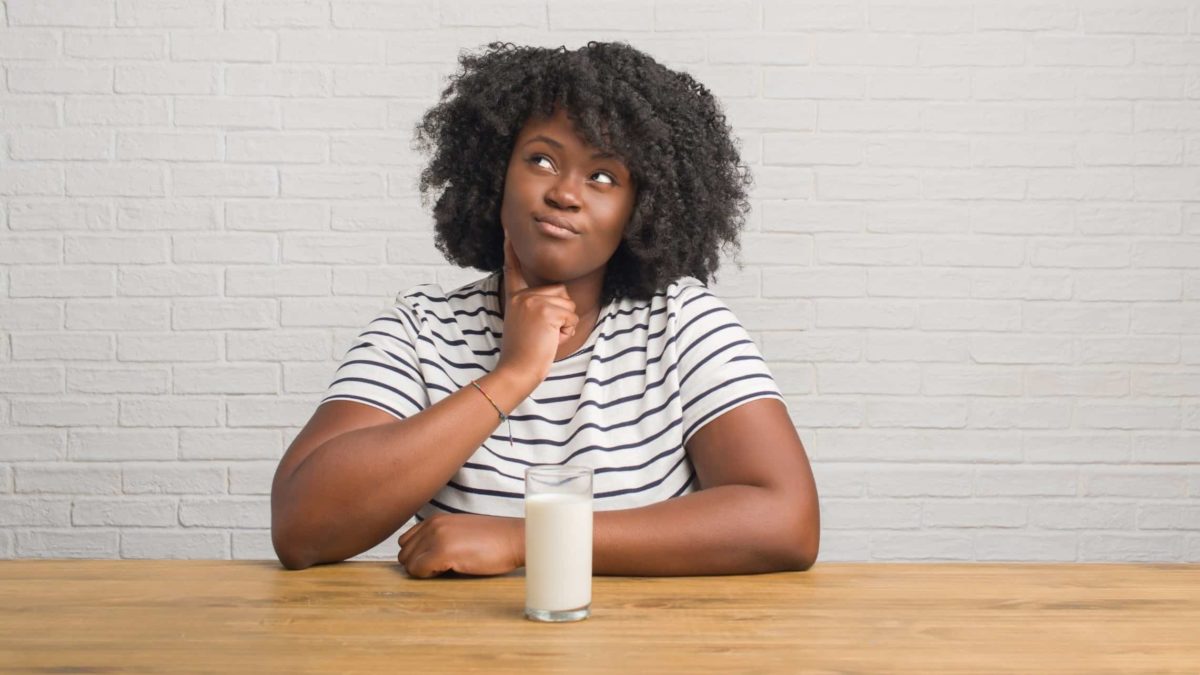 A woman sits with a glass of milk in front of her as she puts a finger to the side of her face as though in thought while her eyes look to the side as though she is contemplating something.