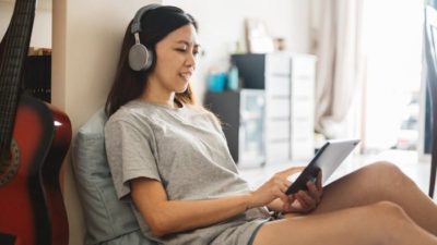 A girl lies on her bed in her room while using laptop and listening to headphones.