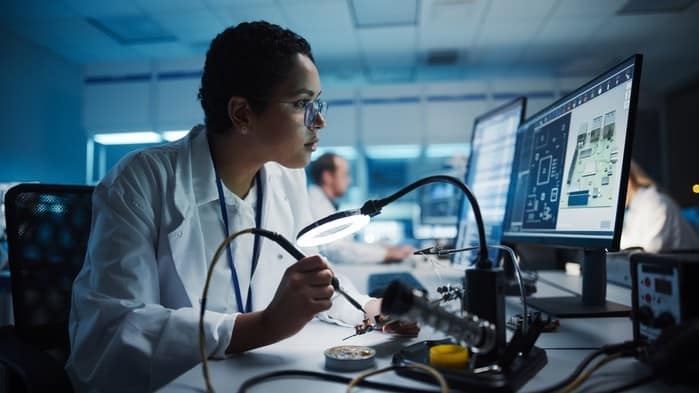 A woman holds a soldering tool as she sits in front of a computer screen while working on the manufacturing of technology equipment in a laboratory environment.