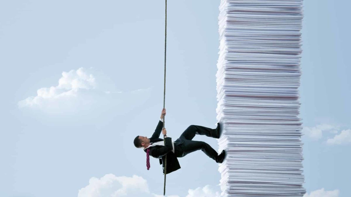 A man in a business suit uses a rope to climb up the side of a huge pile of papers fashioned like a tall building against a blue sky backdrop with clouds representing an assessment of whether CBA shares stacked up well in March